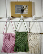 May include: Three macrame wall hangings in pink, green, and cream, suspended from wooden dowels. The hangings are displayed on a white wall. A cardboard box labelled "Make It Yourself Kit Macrame Wall Hanging Pink" is above.