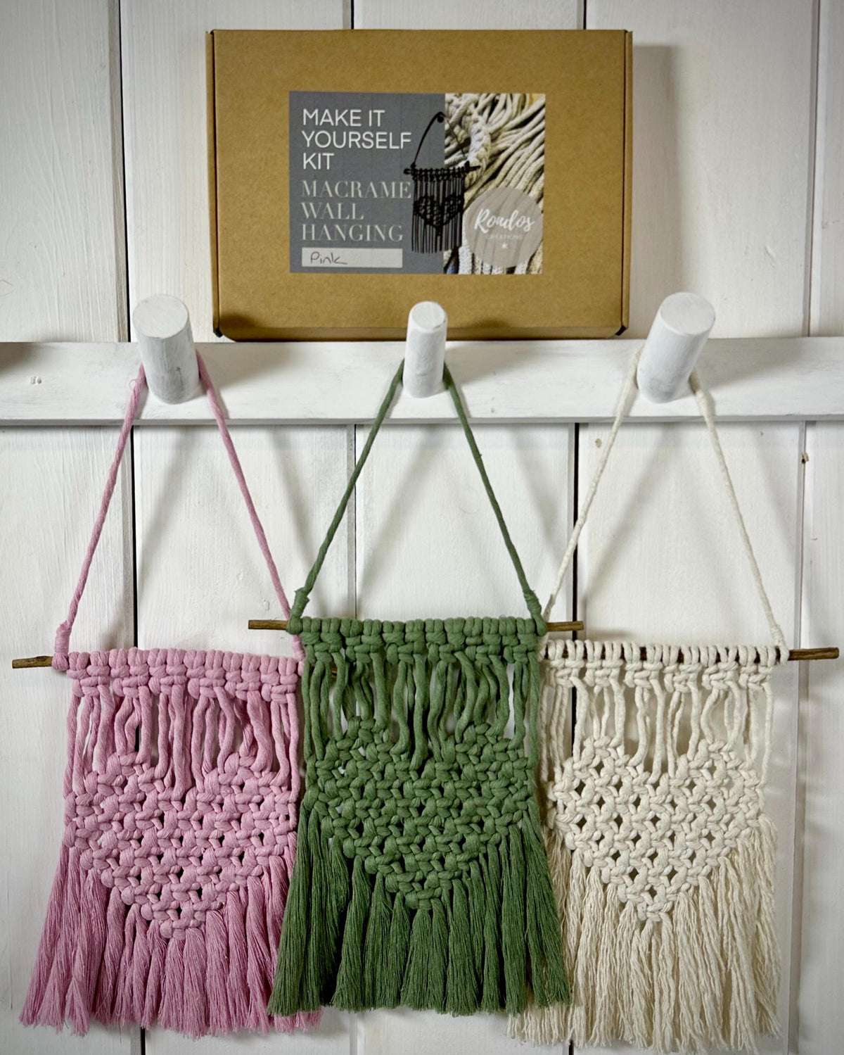 May include: Three macrame wall hangings in pink, green, and cream, suspended from wooden dowels. The hangings are displayed on a white wall. A cardboard box labelled "Make It Yourself Kit Macrame Wall Hanging Pink" is above.